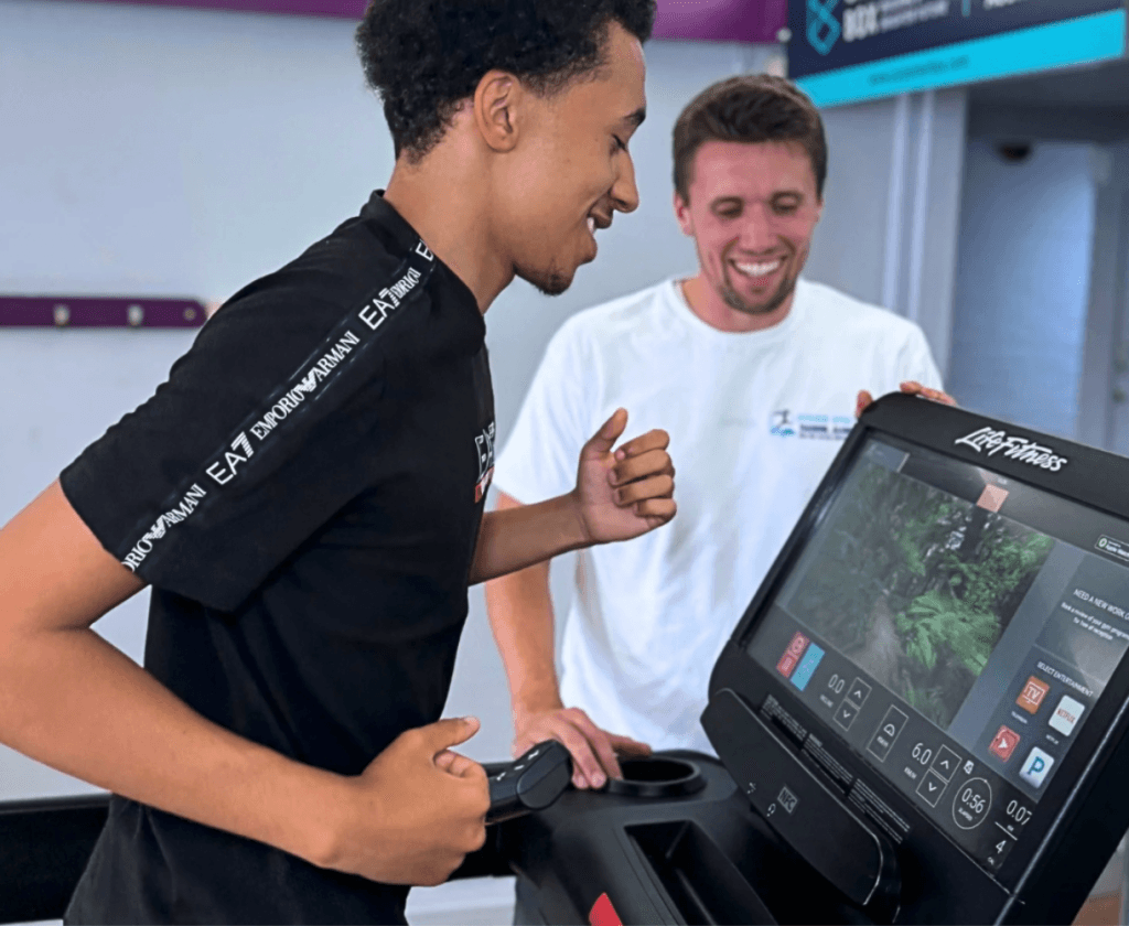 One person runs on a treadmill while another watches beside the console